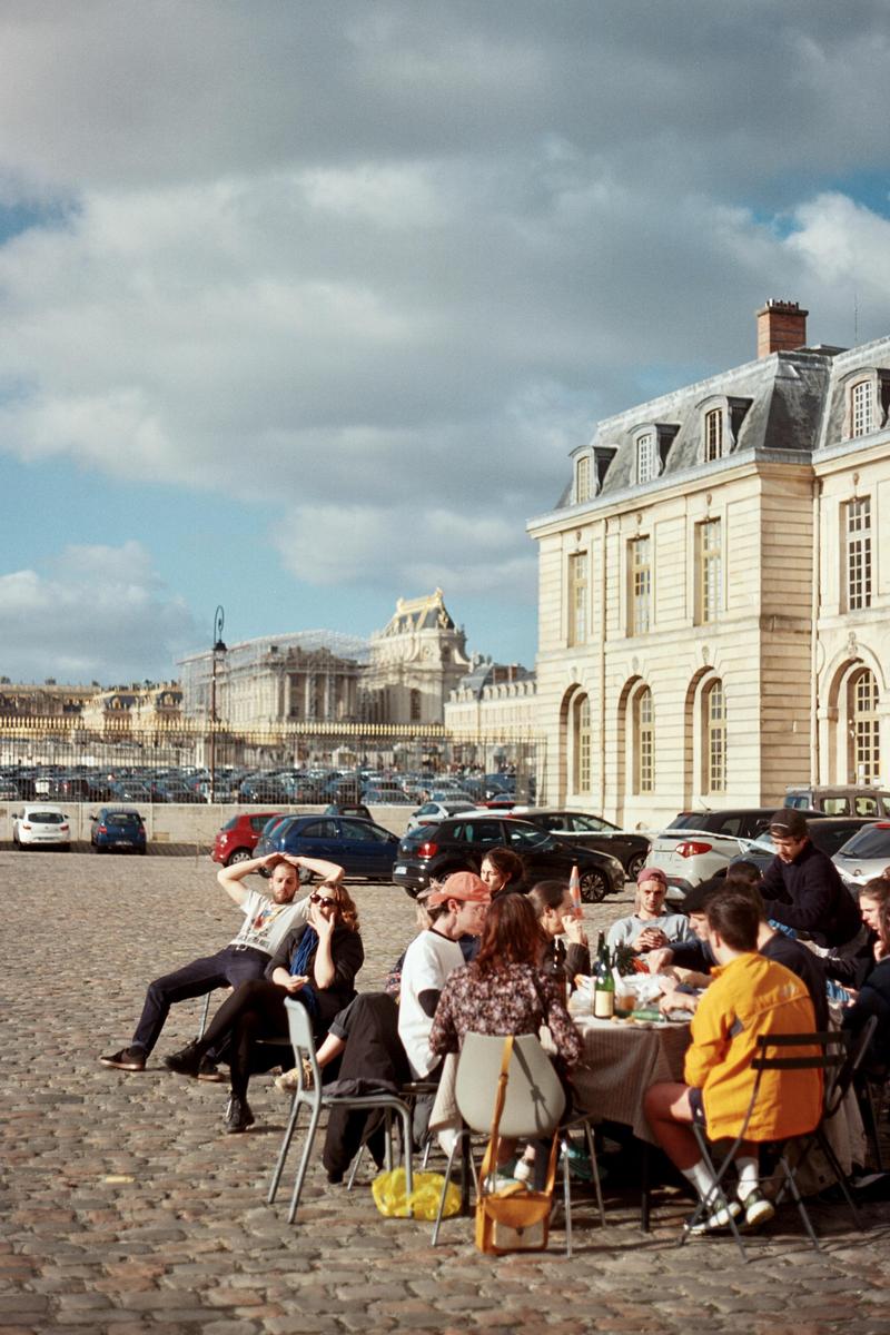 A group of people are seated around a table on a cobblestone street with a large building in the background.