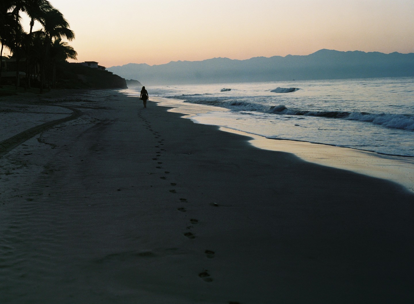 A woman walks along a beach with palm trees and mountains in the distance at dusk.