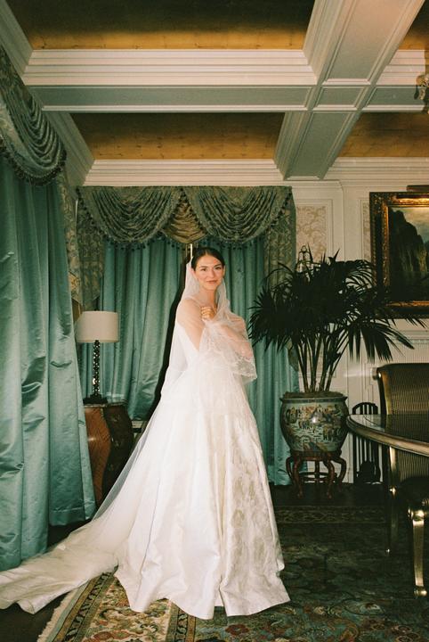 A woman in a wedding dress and veil stands in a room with ornate curtains and a patterned rug.