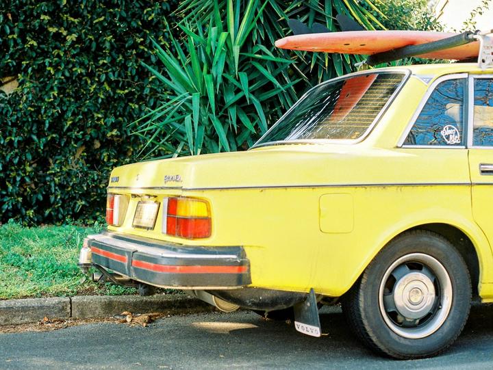 A yellow car with a surfboard on its roof is parked next to a lush green hedge.