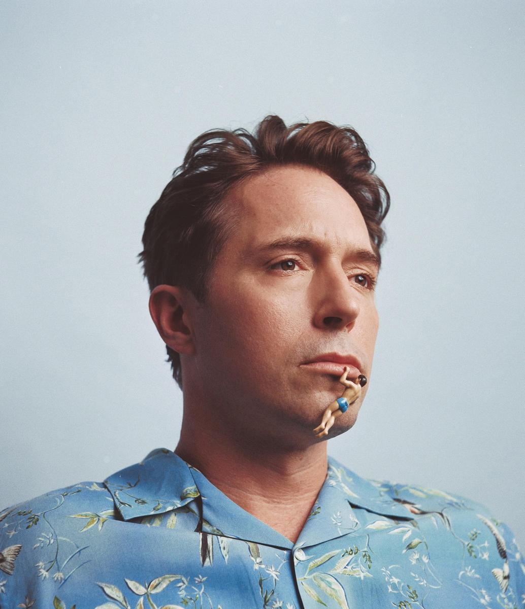A man in a blue patterned shirt has a small figurine of a swimmer hanging from his lower lip.