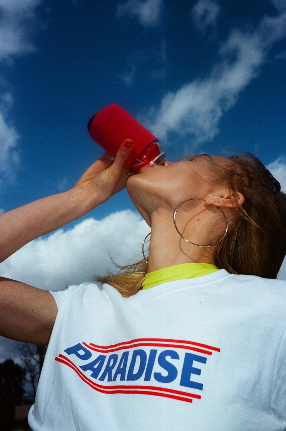 A woman wearing a white t-shirt with the word "paradise" printed on it drinks from a red can against a blue sky with clouds.