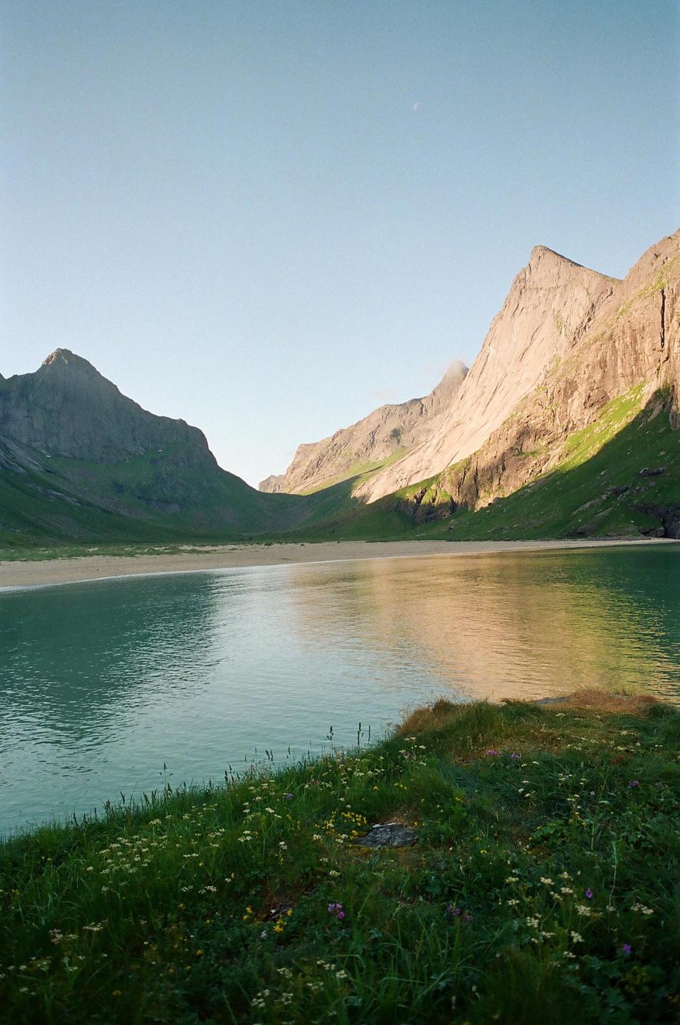 A body of water reflects the sky and mountains, with a sandy beach and grassy bank in the foreground.