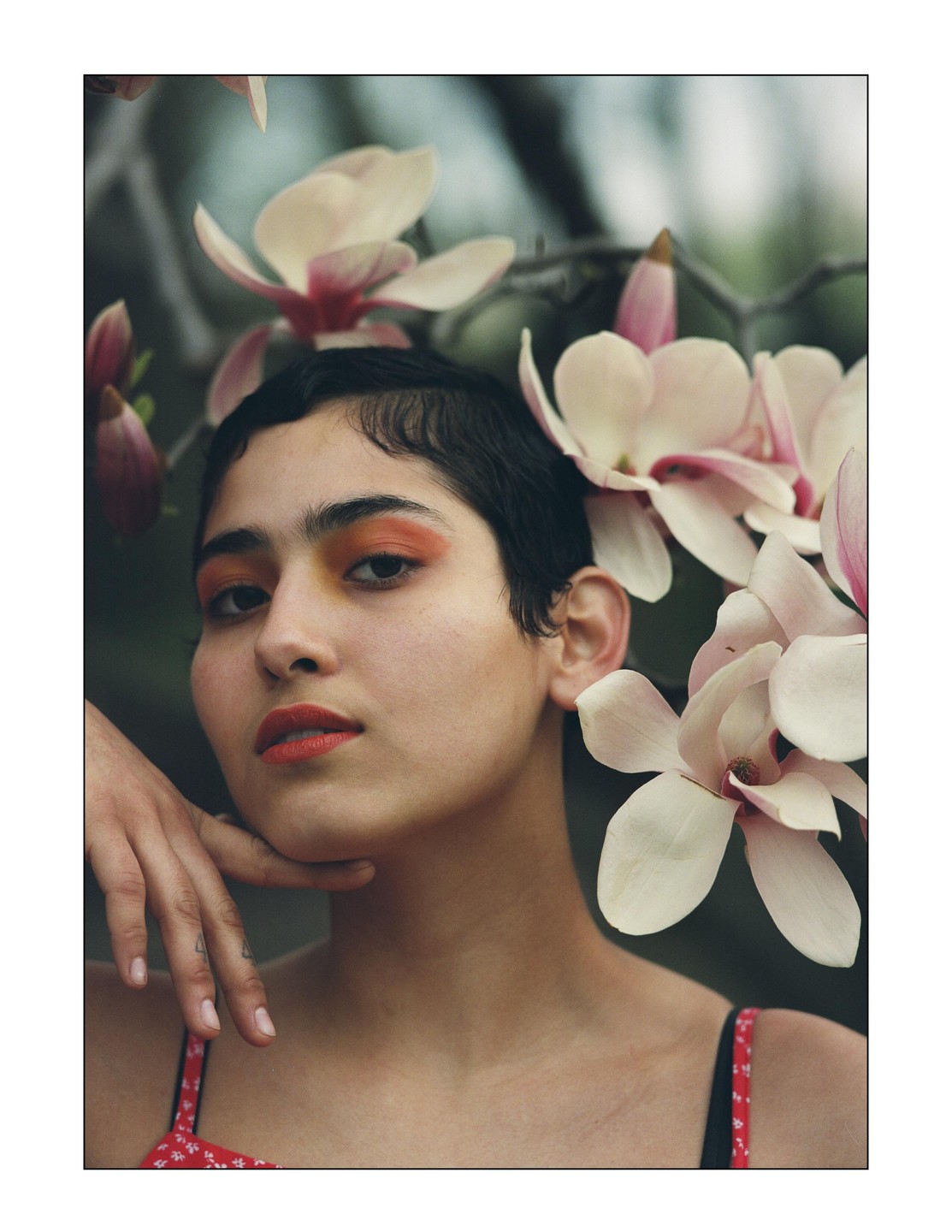 A woman with short hair and orange eyeshadow poses with her hand on her chin, surrounded by magnolia blossoms.