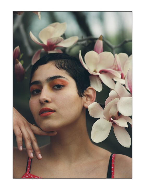 A woman with short hair and orange eyeshadow poses with her hand on her chin, surrounded by magnolia blossoms.