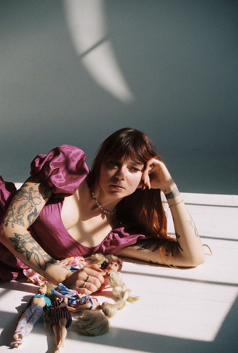 A woman with tattoos and a purple dress is lying on the floor with several dolls.