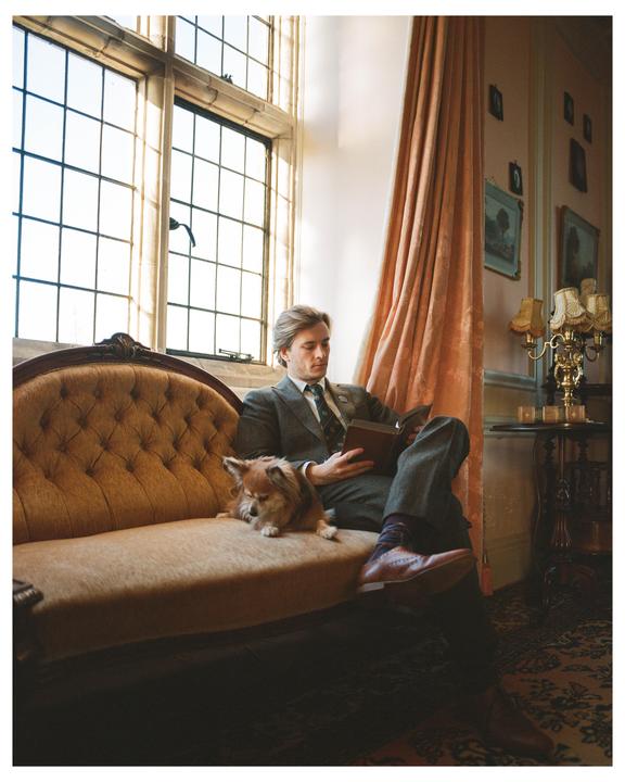 A man in a suit reads a book on a sofa next to a small dog in a room with a large window.