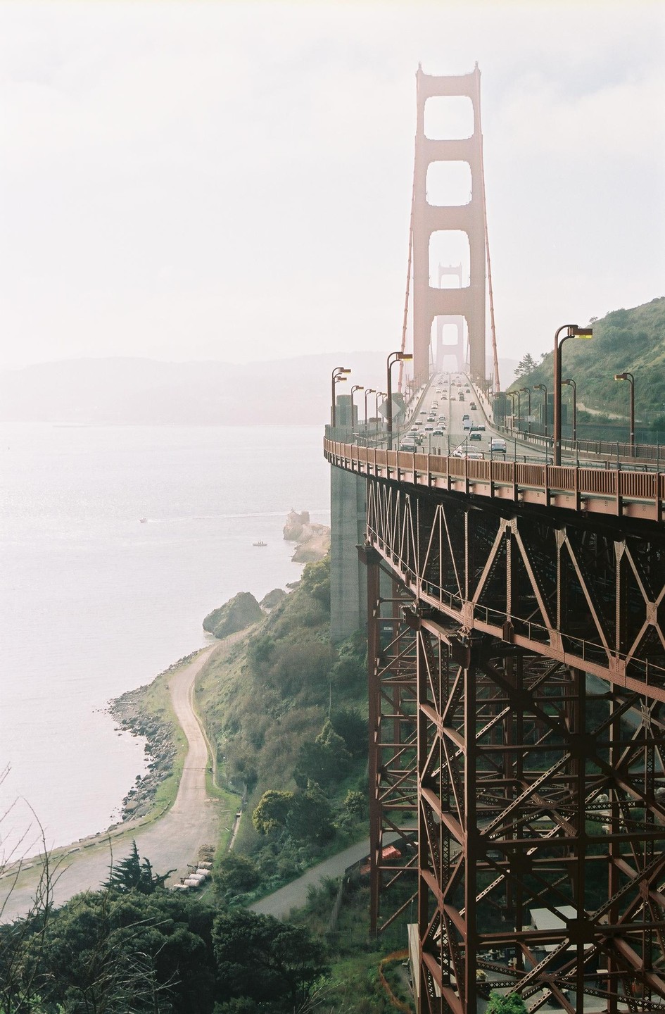 The Golden Gate Bridge stretches across a body of water with a winding road and green hills visible below.