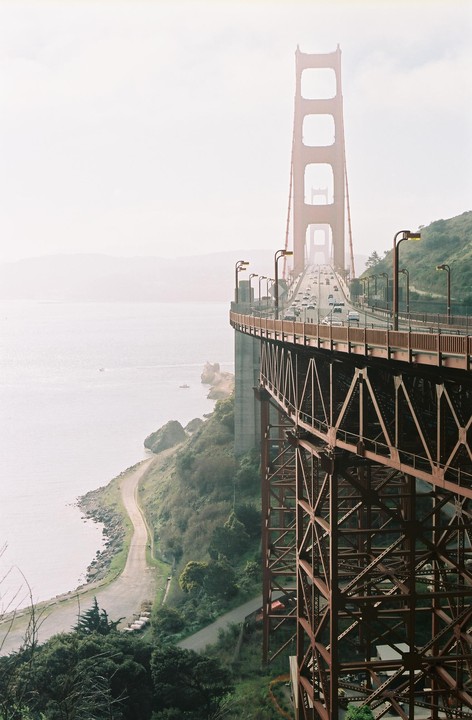 The Golden Gate Bridge stretches across a body of water with a winding road and green hills visible below.