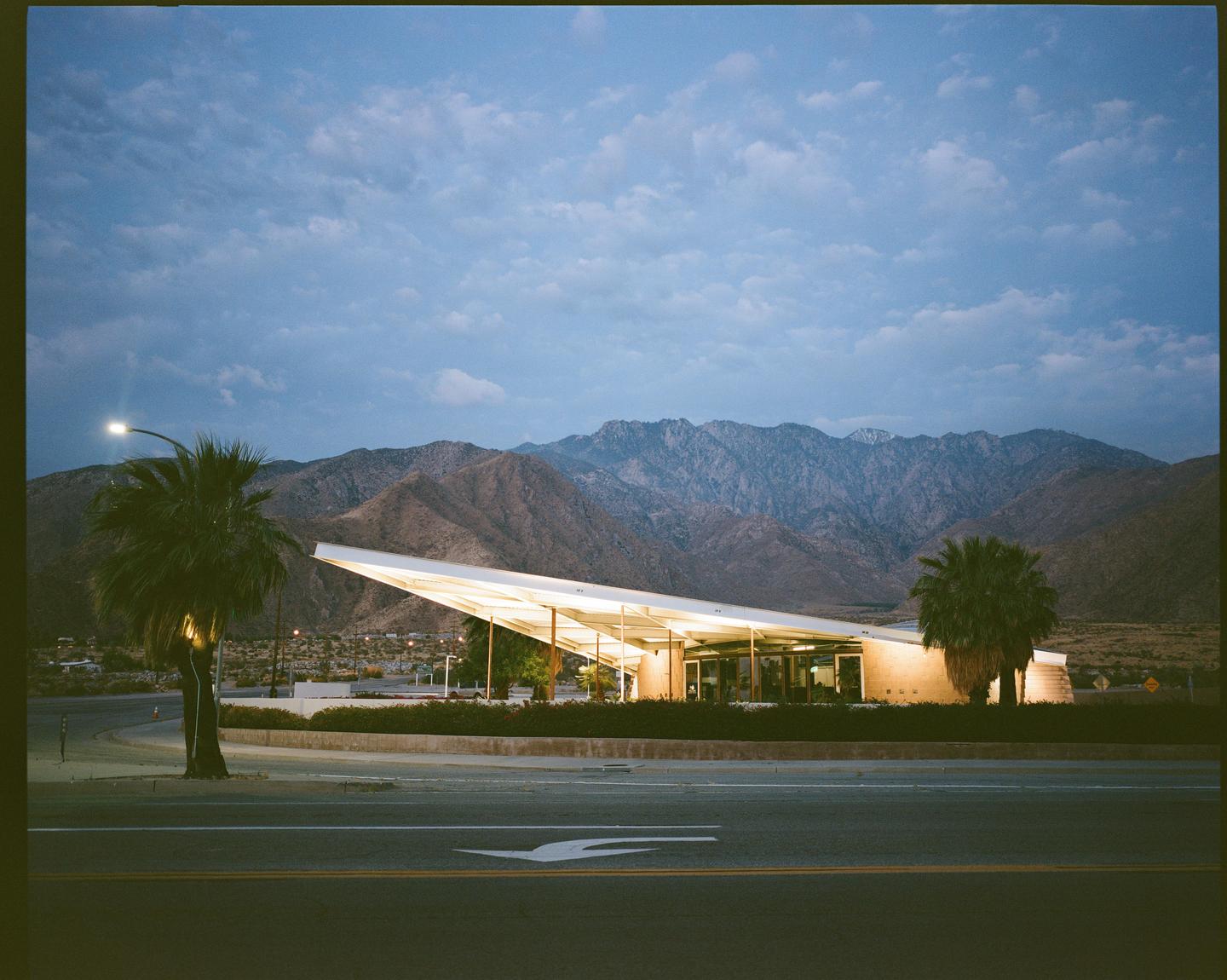 A modern building with a distinctive roofline is illuminated at dusk, with mountains and palm trees in the background.