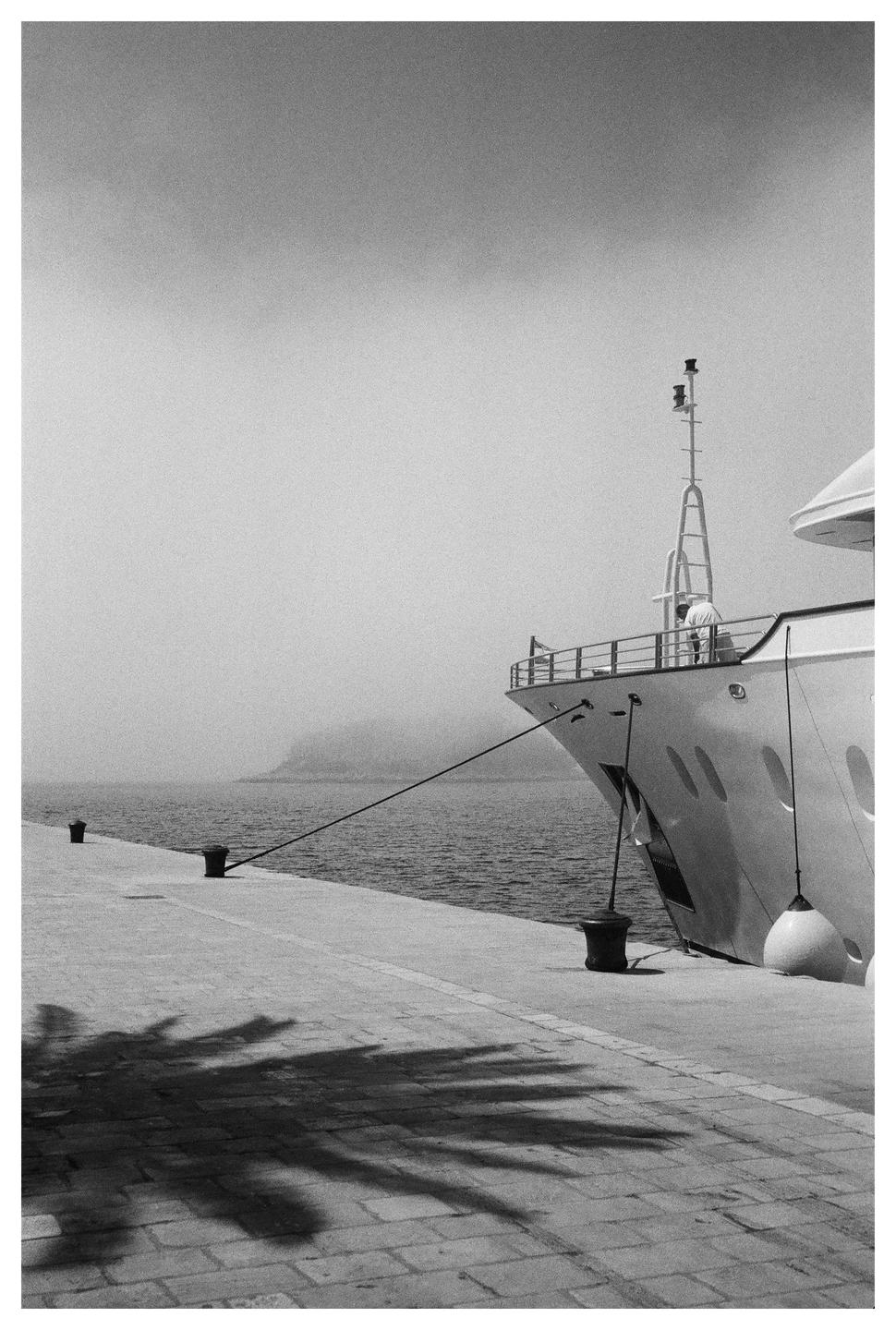 A large yacht is docked at a pier with palm tree shadows in the foreground and a hazy mountain in the distance.