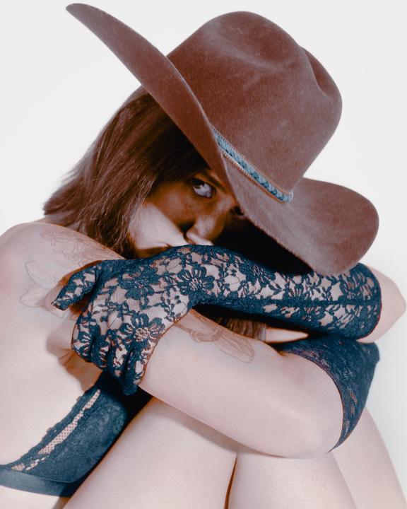 A woman wearing a cowboy hat, lace gloves, and underwear is looking at the viewer.