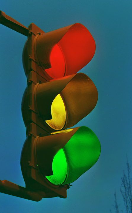 A traffic light with red, yellow, and green lights illuminated is viewed from below against a blue sky.