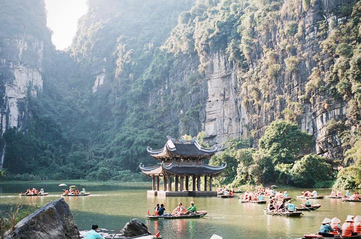 A pagoda stands in a lake surrounded by boats with people and steep, tree-covered cliffs.