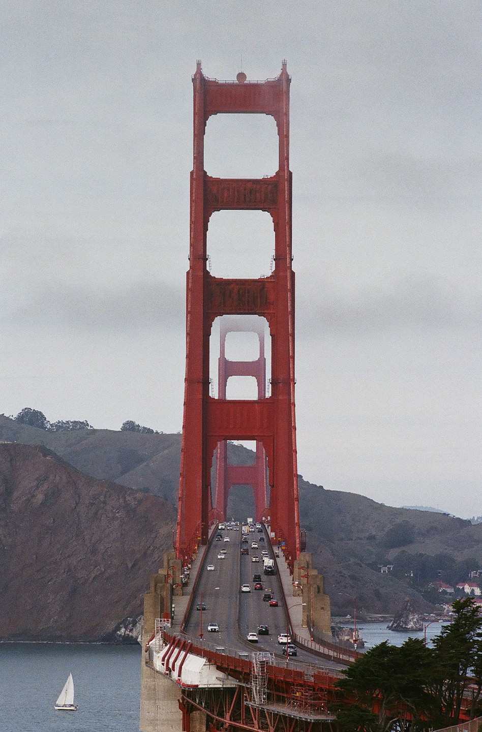 The Golden Gate Bridge is visible from a distance, with a sailboat on the water and cars on the bridge.