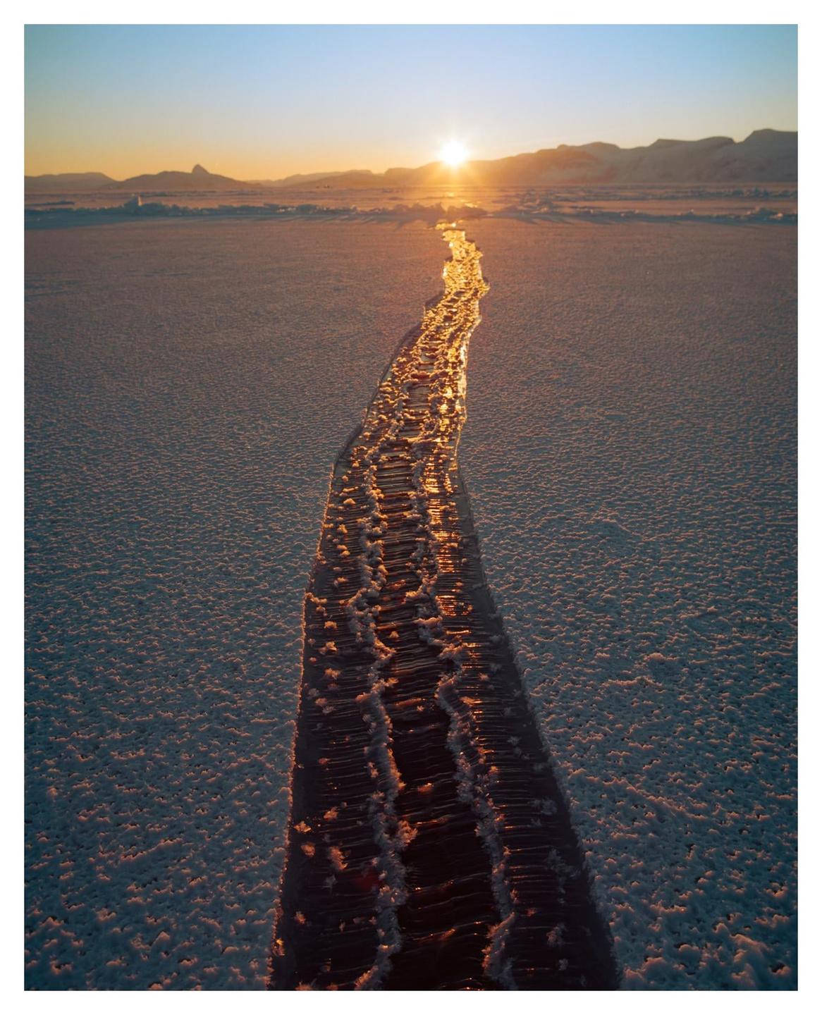 A crack in an icy landscape reflects the golden light of the setting sun.