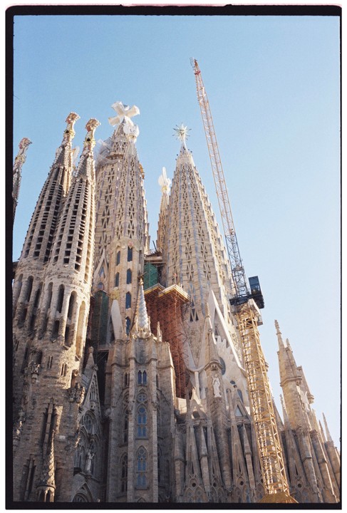 A low-angle view shows the Sagrada Familia cathedral under construction against a clear sky.