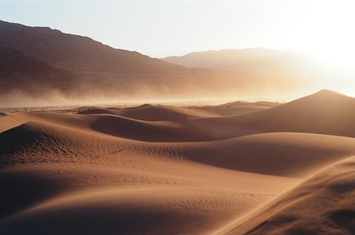 A desert landscape shows sand dunes with mountains in the background under a hazy sky.