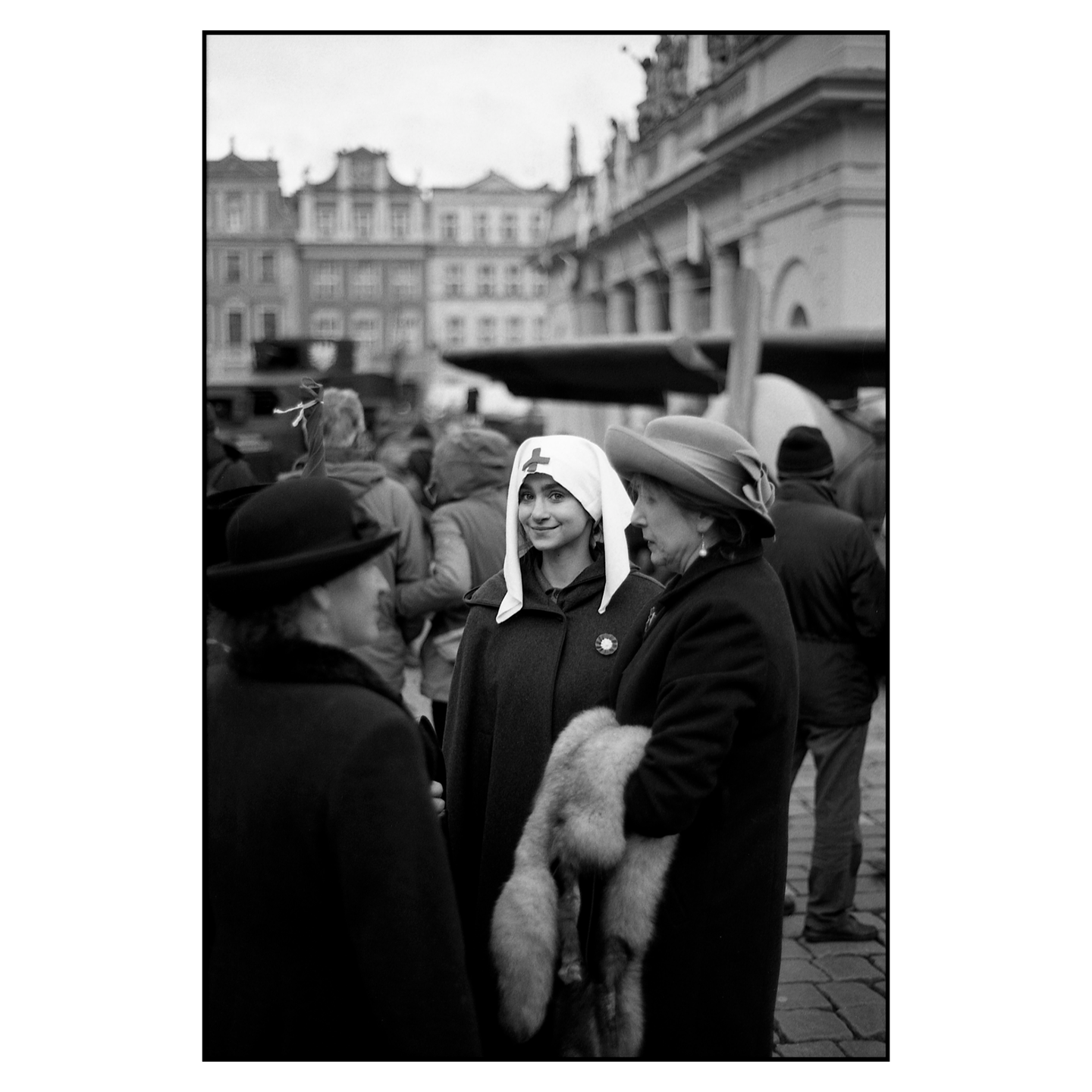 Three women stand on a cobblestone street, one wearing a white head covering with a red cross, another a hat and fur stole, and the third a hat.
