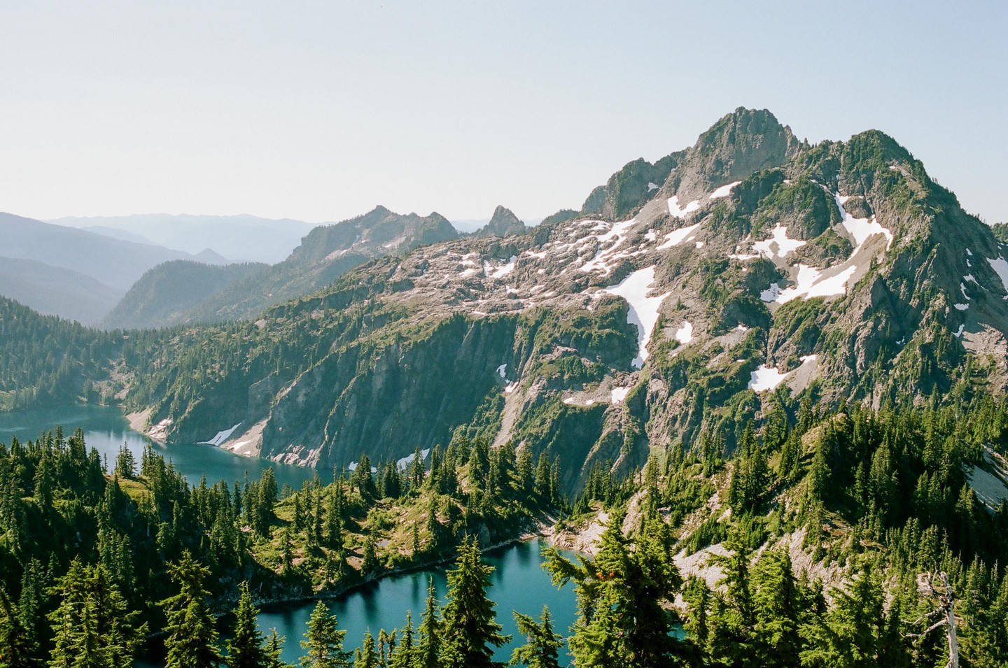 A high-angle landscape shows a mountain lake surrounded by pine trees and snow-capped peaks under a clear sky.