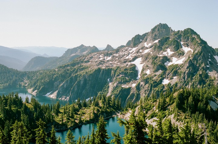 A high-angle landscape shows a mountain lake surrounded by pine trees and snow-capped peaks under a clear sky.