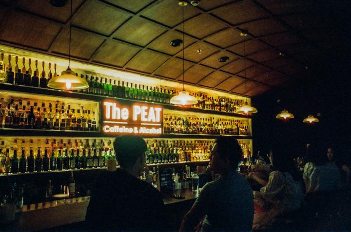 People sit at a bar with a large display of liquor bottles and a neon sign that reads "The PEAT Caffeine & Alcohol."