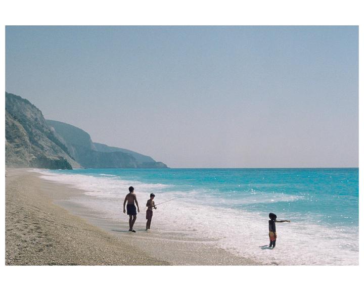 A man, a boy, and a child are on a beach with cliffs in the background and turquoise water.