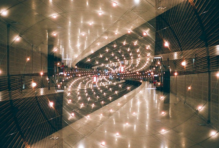 A double exposure shows the interior of a train station with many lights reflecting on the floor and ceiling.