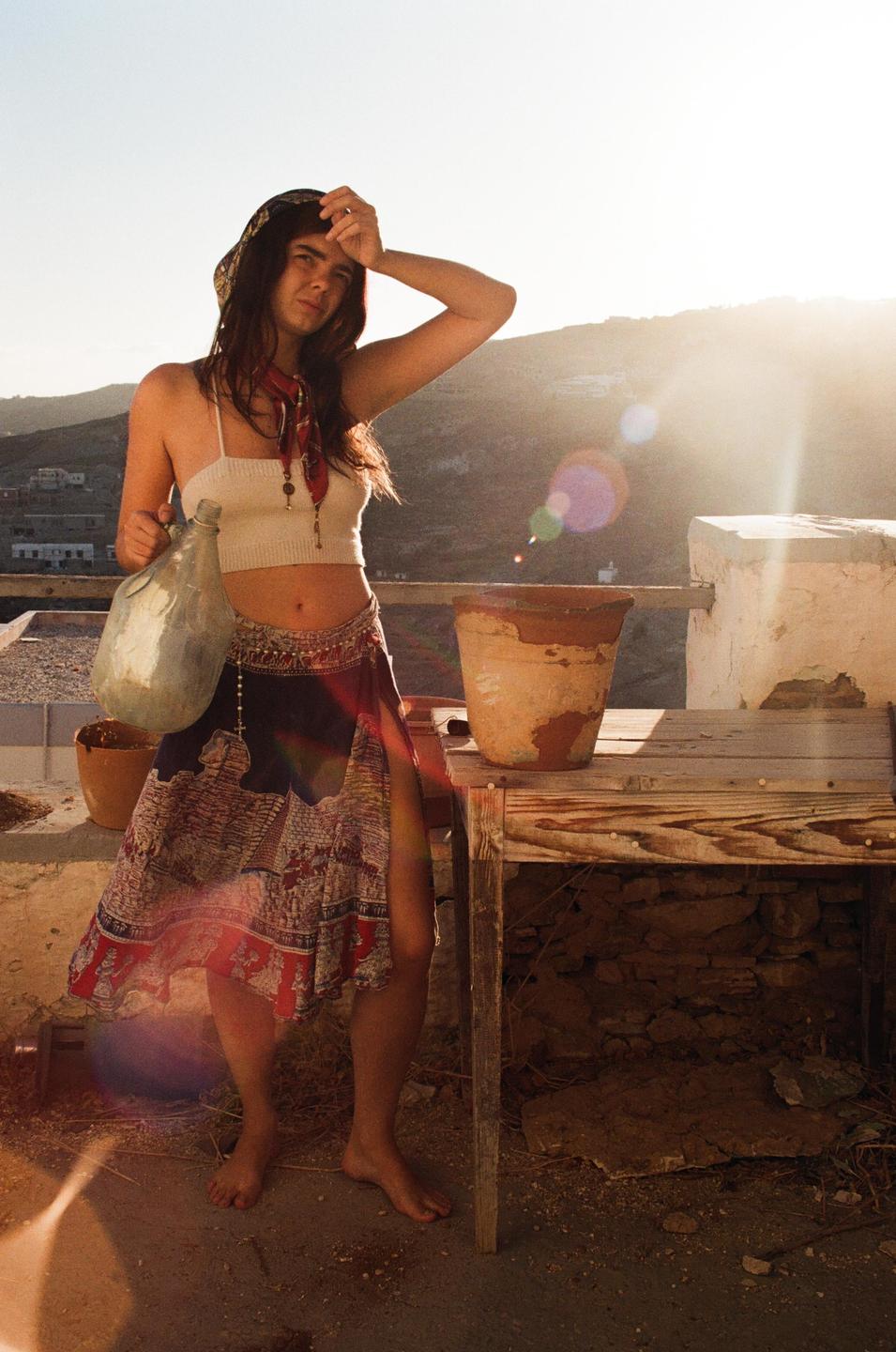 A woman stands barefoot on a patio, holding a large glass bottle, with mountains and a bright sky in the background.