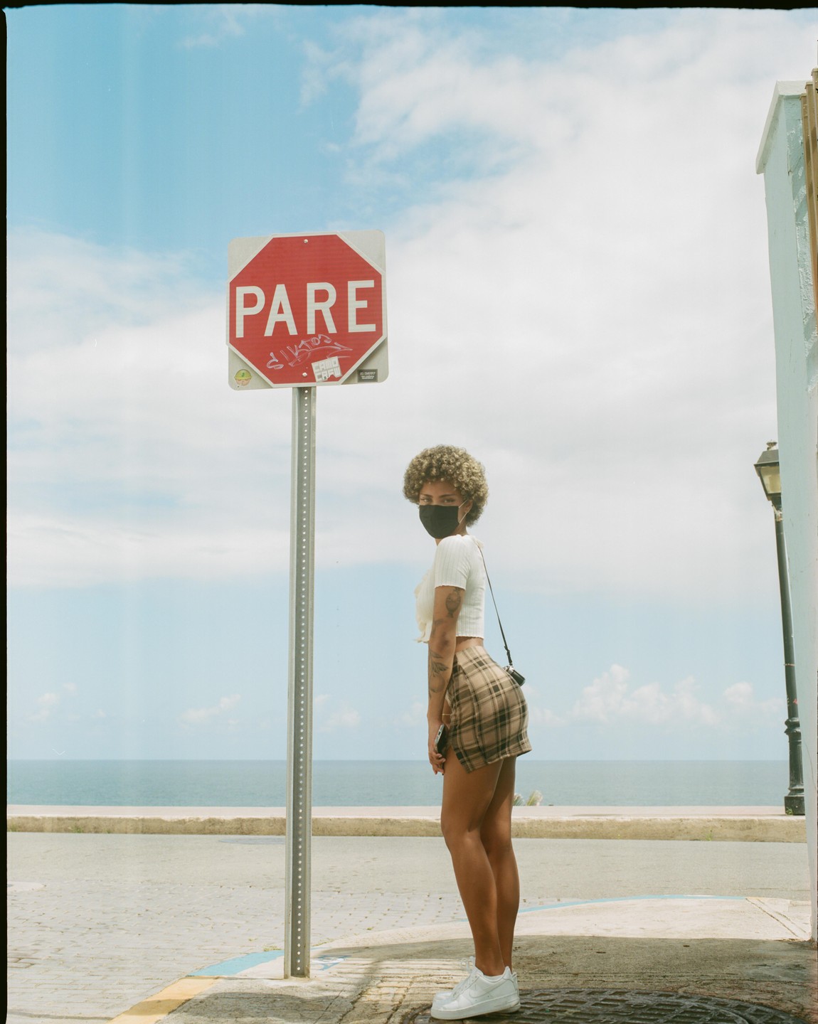 A woman in a plaid skirt and face mask stands next to a stop sign with the ocean in the background.