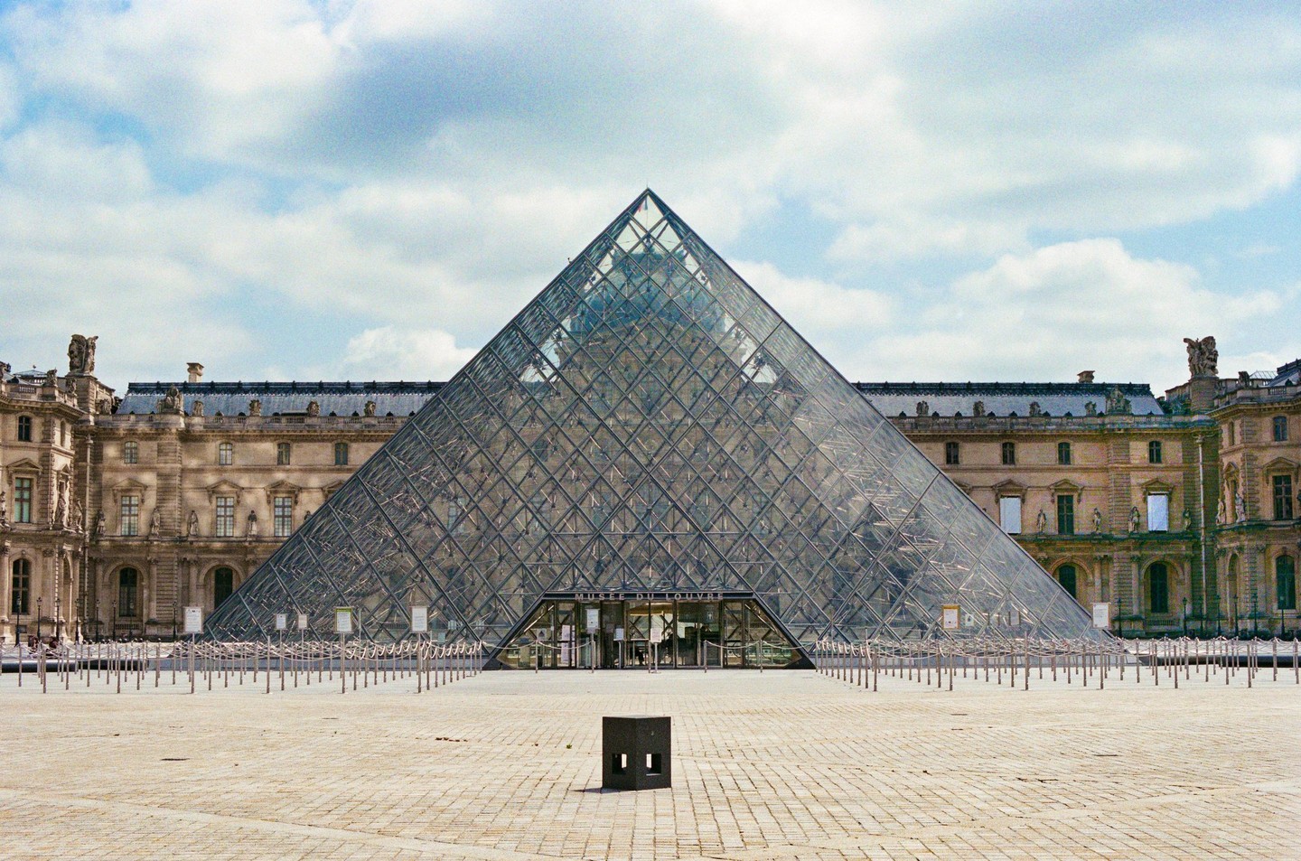 The Louvre Pyramid stands in front of the historic palace under a cloudy sky.