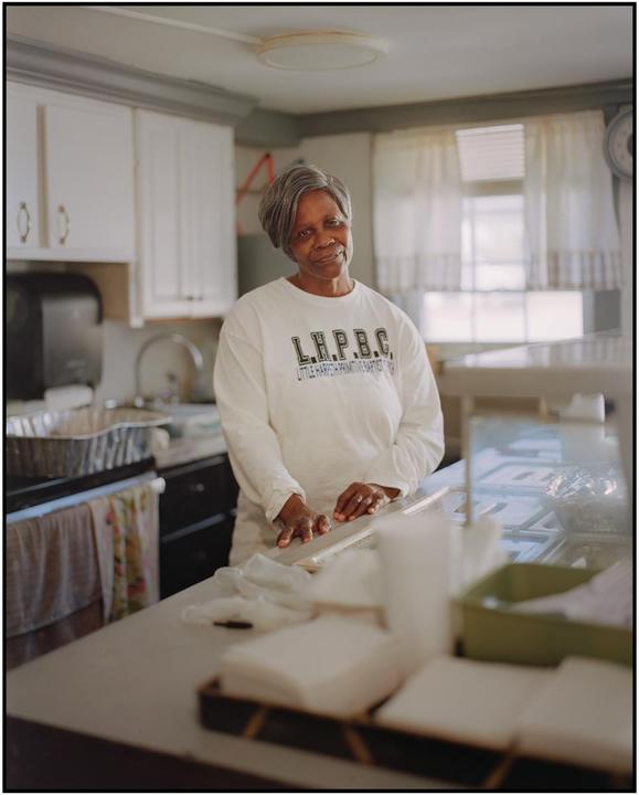 A woman stands in a kitchen with white cabinets and a window with white curtains.