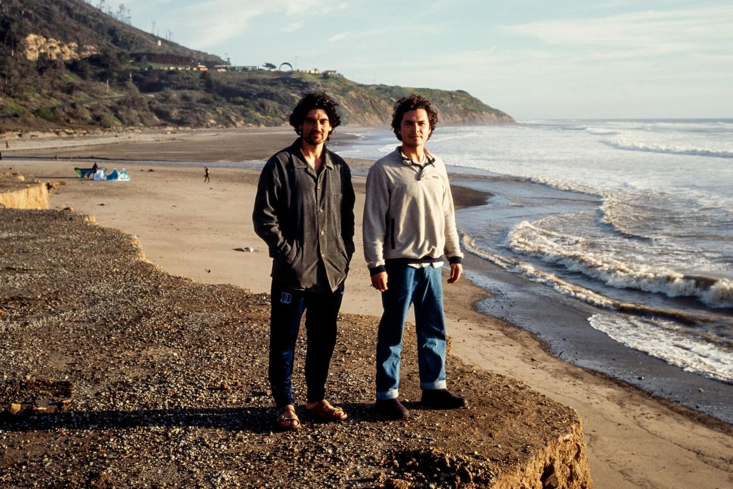 Two men stand on a cliff overlooking a beach with waves crashing on the shore.