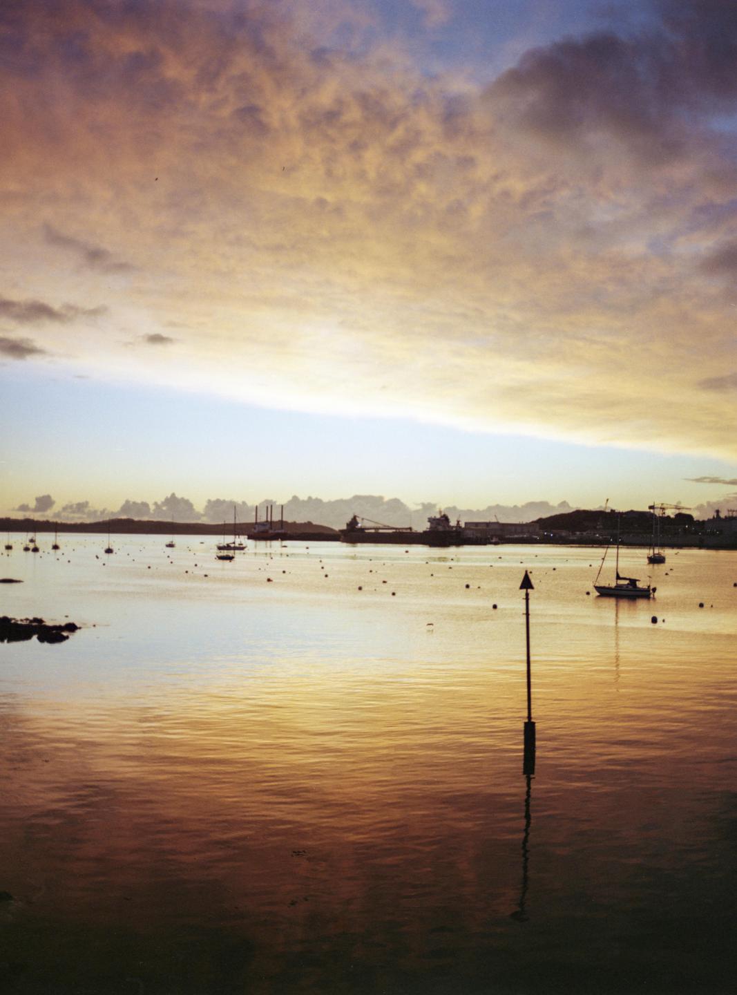 A body of water with several boats and buoys is visible under a sky with golden clouds.