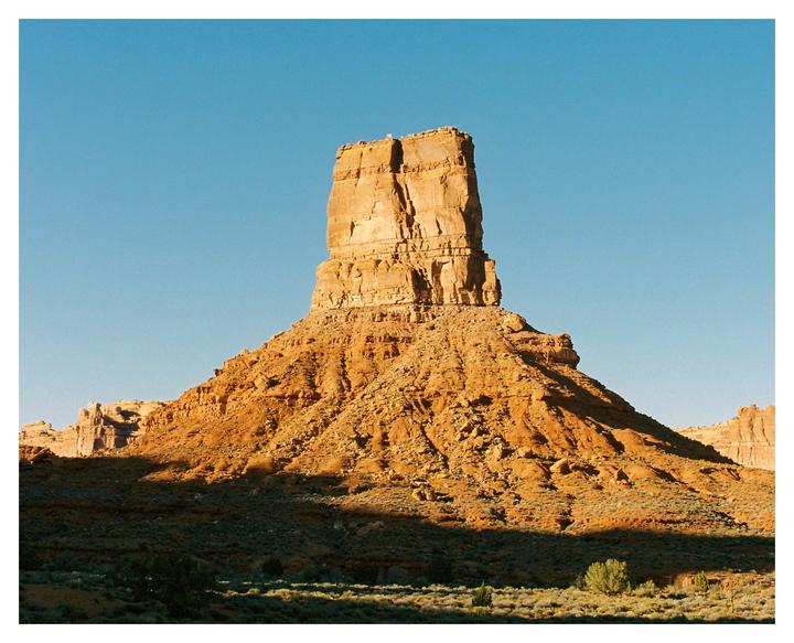 A large rock formation stands tall against a clear blue sky in a desert landscape.