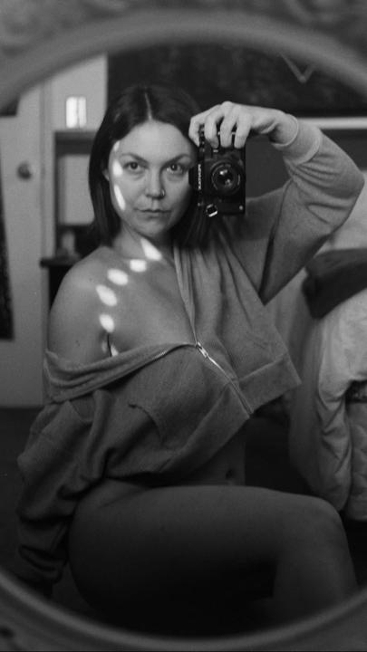 A woman takes a self-portrait in a mirror, holding a camera to her face, with dappled light on her shoulder.