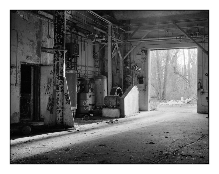 Sunlight streams into an abandoned industrial building through a large open doorway, revealing graffiti-covered walls and machinery.