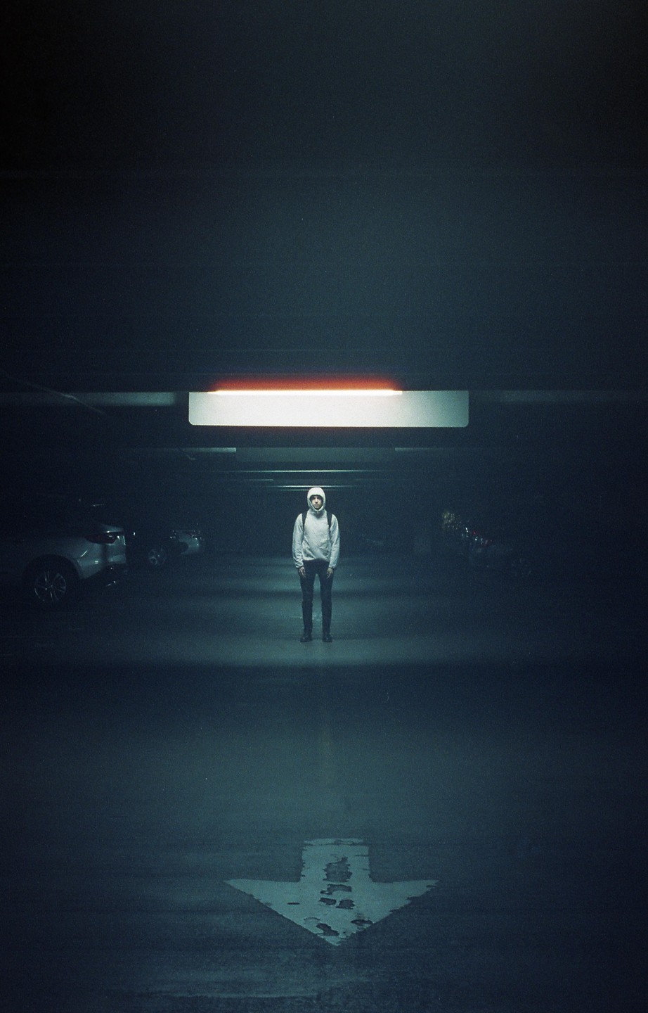 A person wearing a hoodie and backpack stands in a dimly lit parking garage with an arrow painted on the ground.