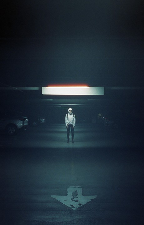 A person wearing a hoodie and backpack stands in a dimly lit parking garage with an arrow painted on the ground.