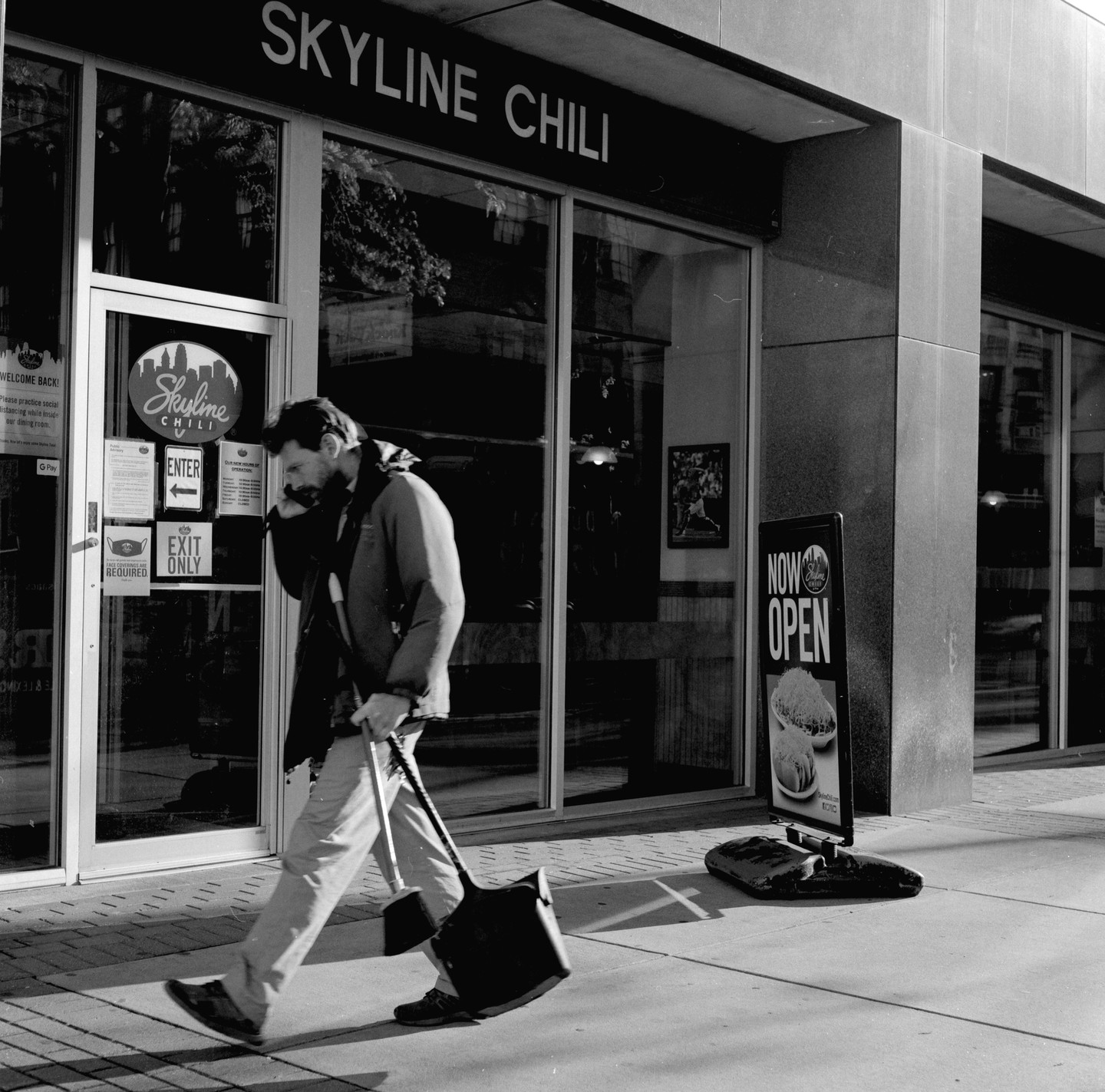 A man walks past a Skyline Chili restaurant while talking on a phone and carrying a shovel.