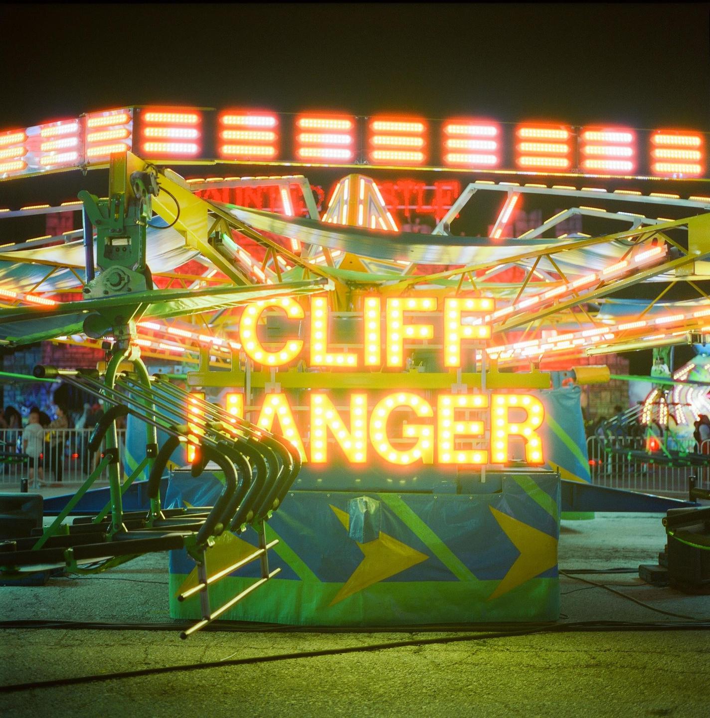 A brightly lit carnival ride with the words "CLIFF HANGER" in glowing yellow letters is visible at night.