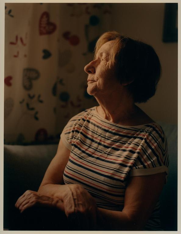 A woman with her eyes closed is illuminated by a ray of light, sitting in front of a curtain with heart patterns.