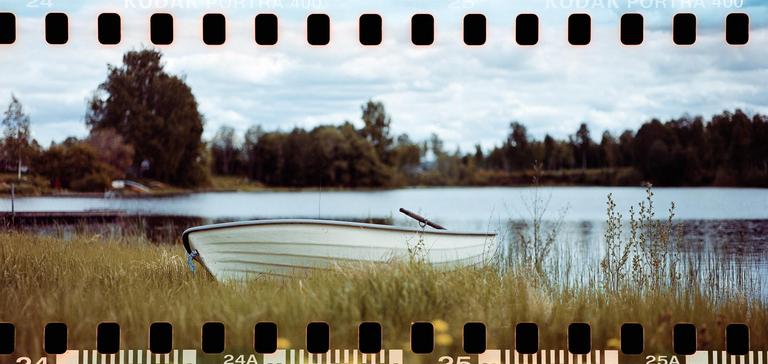 A small boat rests on a grassy bank beside a lake with trees in the background.