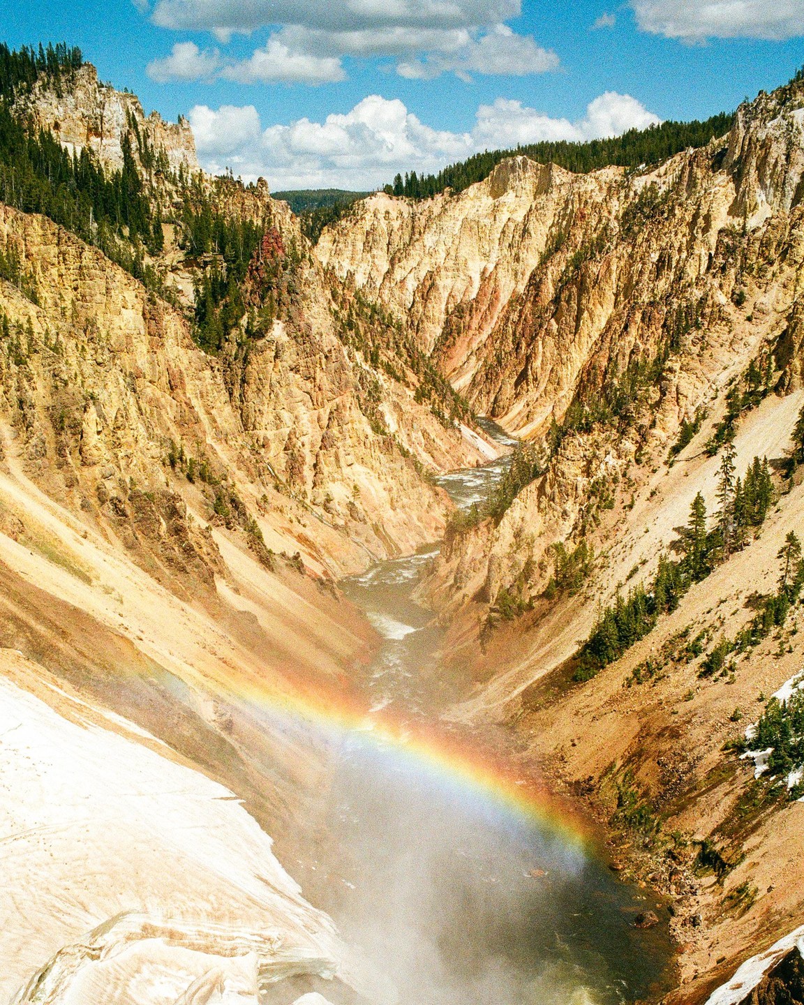 A rainbow stretches across a canyon with a river flowing through it, surrounded by rocky cliffs and green trees.