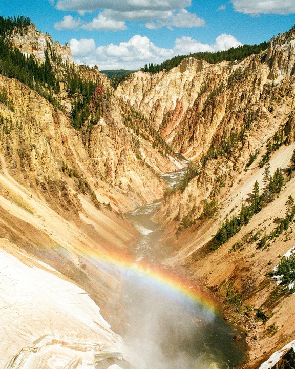 A rainbow stretches across a canyon with a river flowing through it, surrounded by rocky cliffs and green trees.