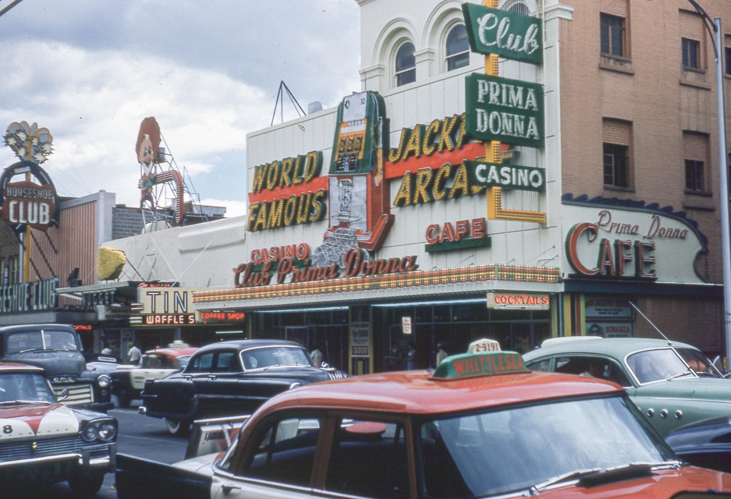 Cars are parked on a street in front of a casino and cafe with large neon signs.