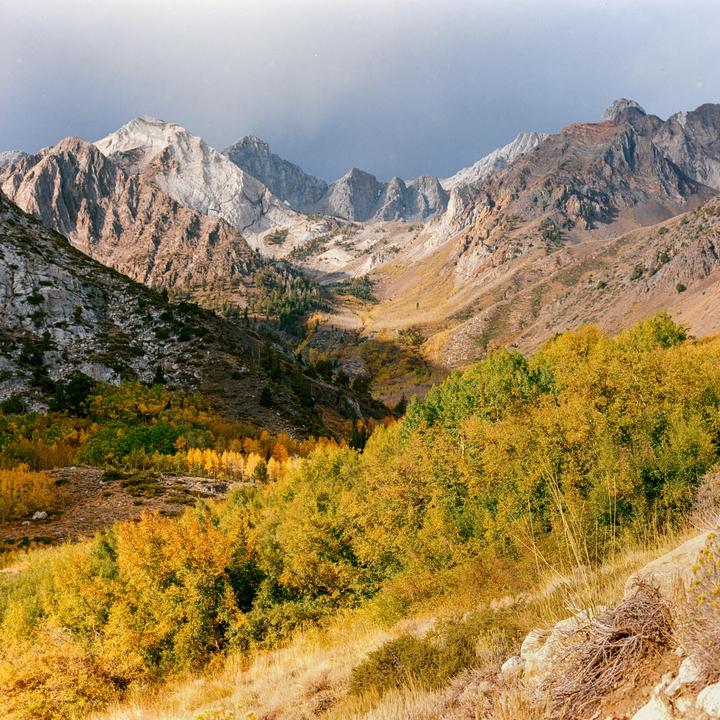 A valley with autumn foliage is nestled between rocky mountains under a cloudy sky.