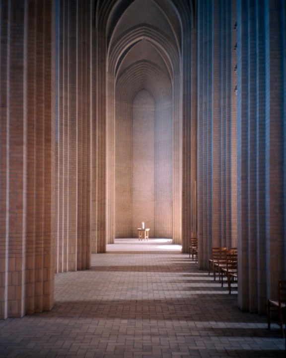 A long hall with tall columns and arched ceilings leads to a small table with a candle.