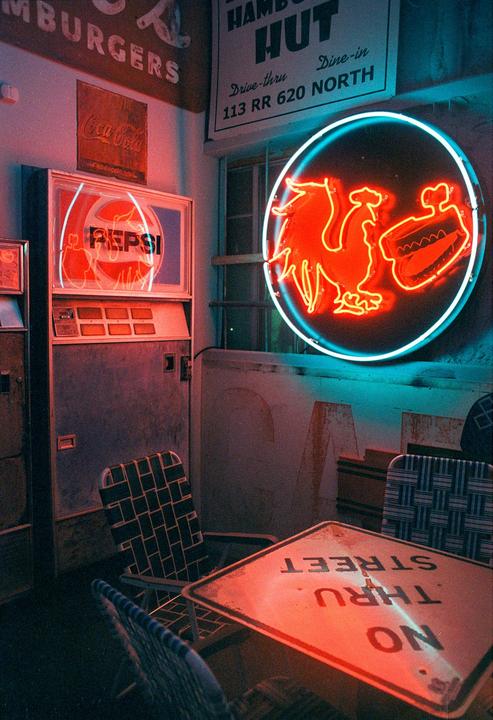 A neon rooster and hot dog sign glows above a table with a "no thru street" sign on it, next to a Pepsi machine.