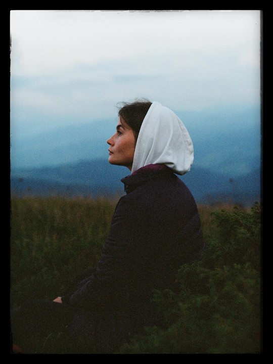 A woman wearing a white head covering sits in a field with mountains in the background.